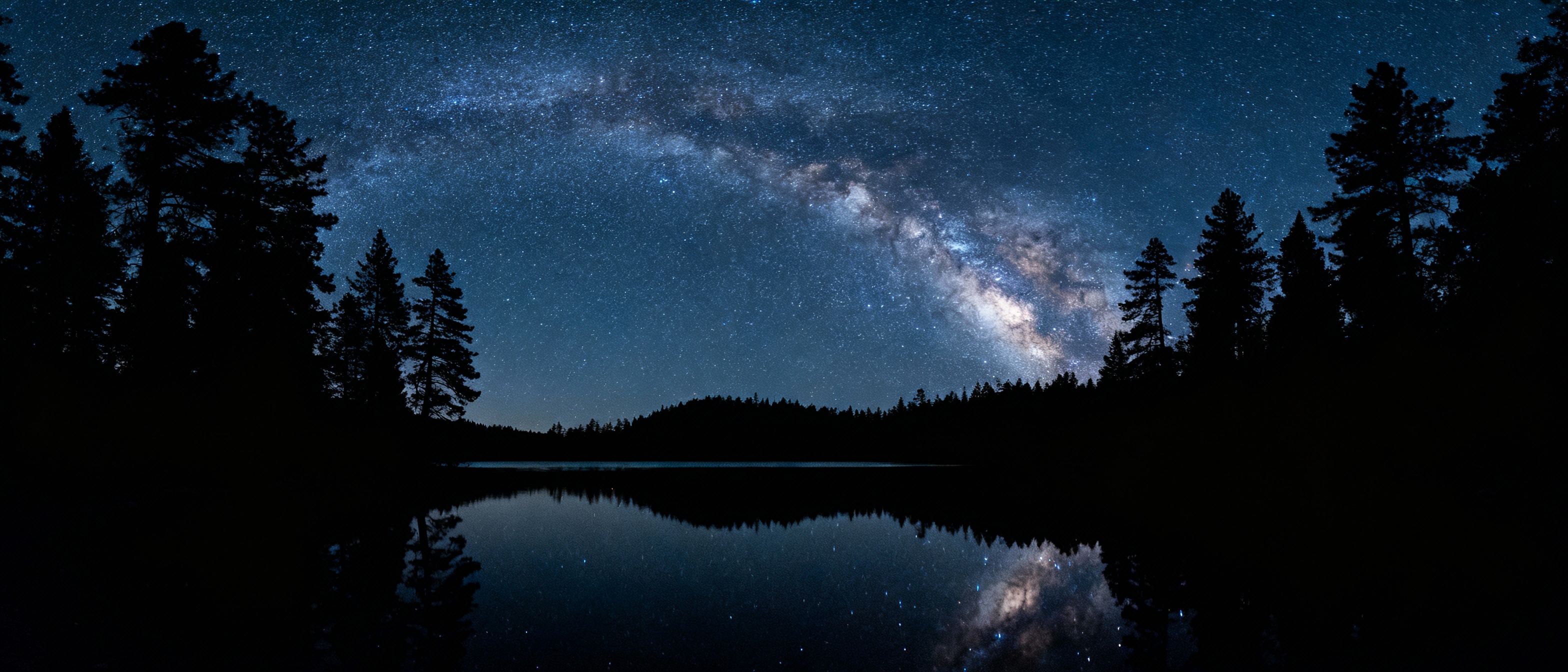 A starry sky with the Milky Way arching above a serene lake, captured using a wide-angle prime lens at f/1.8. The foreground water reflects the galaxy’s core, rendered in crisp detail with minimal noise. Dark silhouettes of pine trees anchor the scene, enhanced by the lens’s edge-to-edge sharpness.