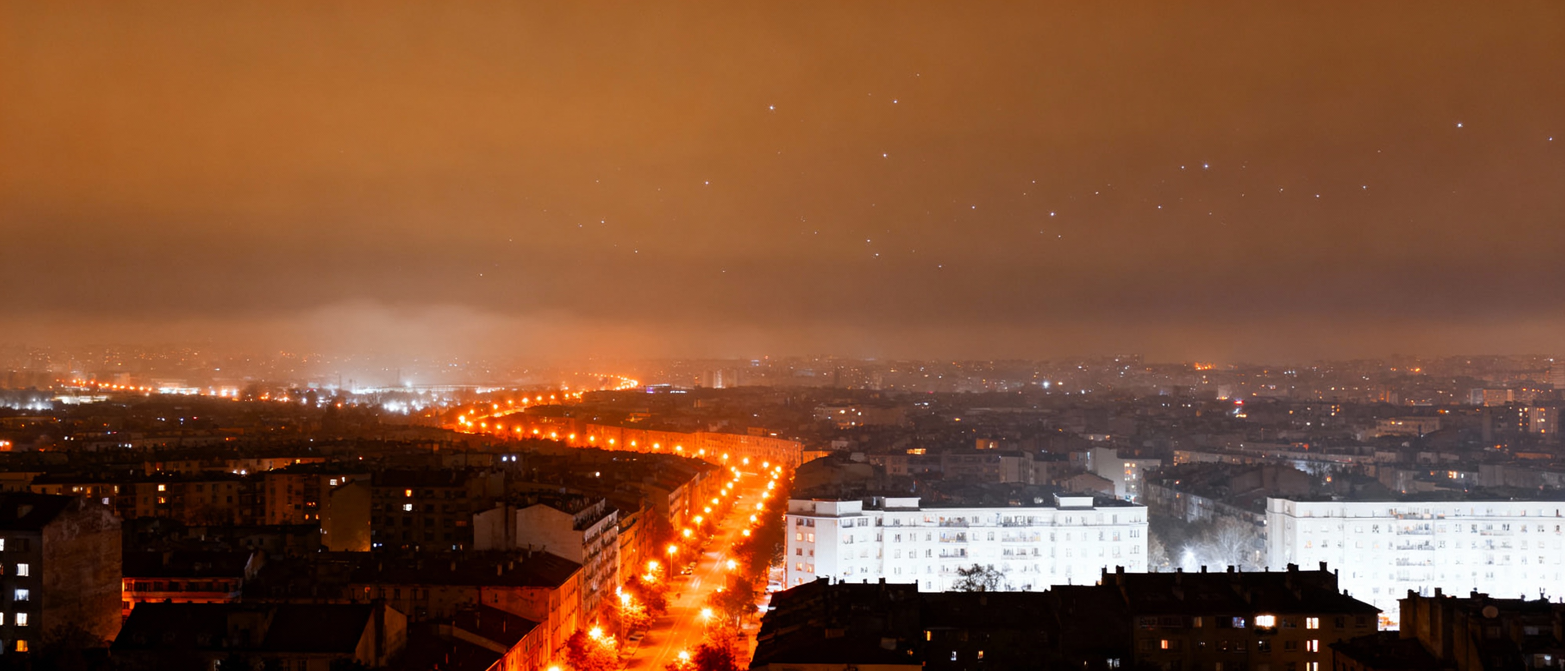 A sprawling cityscape at night illuminated by orange and white streetlights, with a hazy sky glowing amber above, contrasting sharply against a distant horizon where faint stars struggle to pierce through light pollution.